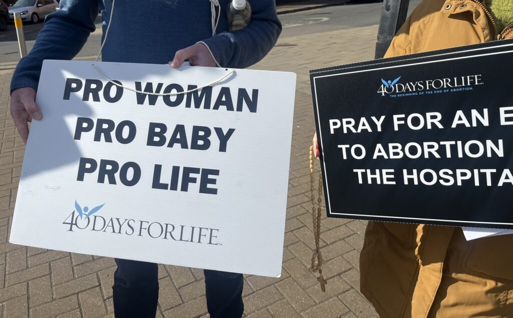 ALT TEXT: Two people holding signs, one with a white sign and a 40 Days to Life logo reading “Pro woman, pro baby, pro life” and one black sign reading “Pray for an end to abortion in the hospital”