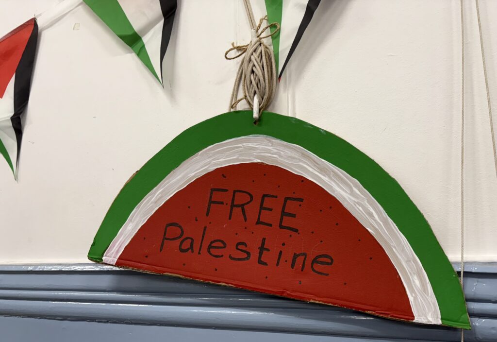 A carboard sign, painted and cut to appear as a watermelon, that reads Free Palestine. Hung on a white wall. 