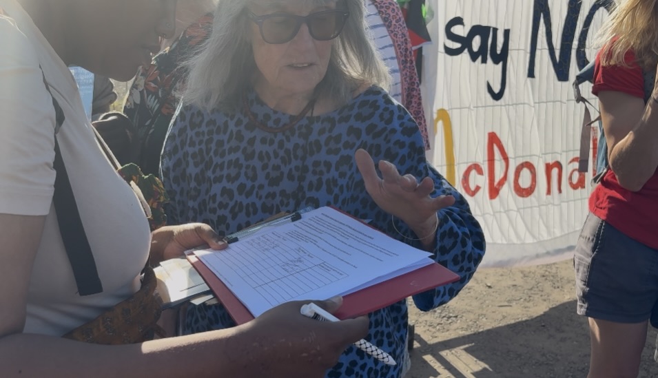 Woman in sunglasses holding a red clipboard while another person signs a petition, with a protest banner in the background.