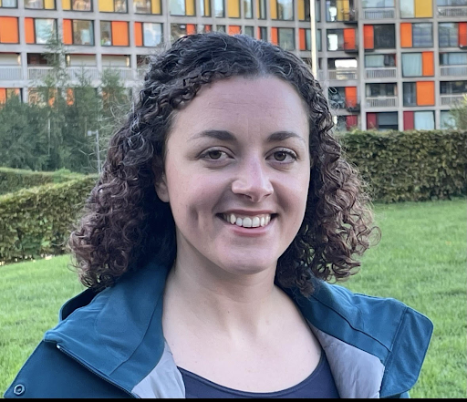Woman pictured in front of a block of flats smiling with a blue coat on.