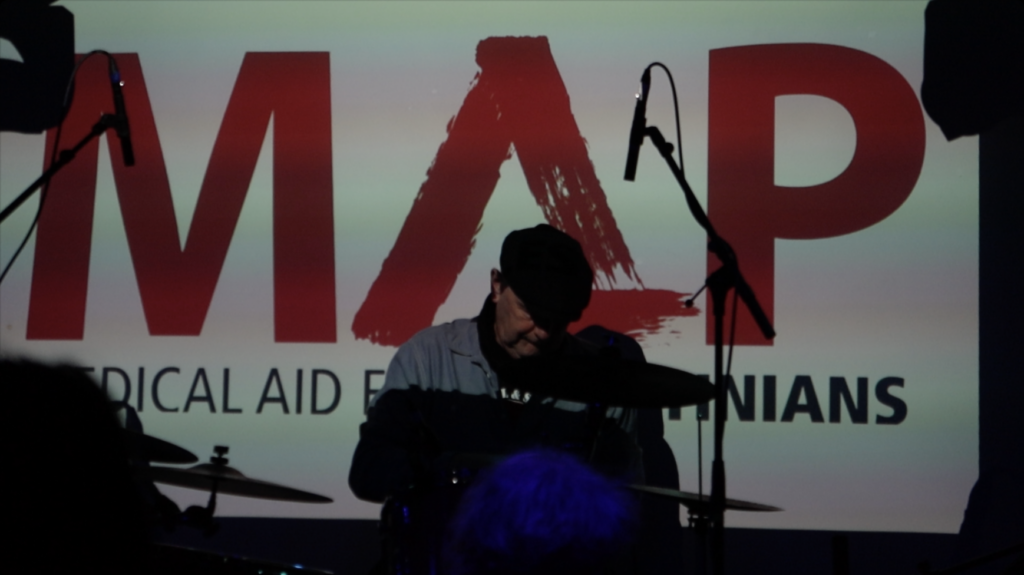 Blues drummer on stage playing with the Medical Aid for Palestinians logo in the background