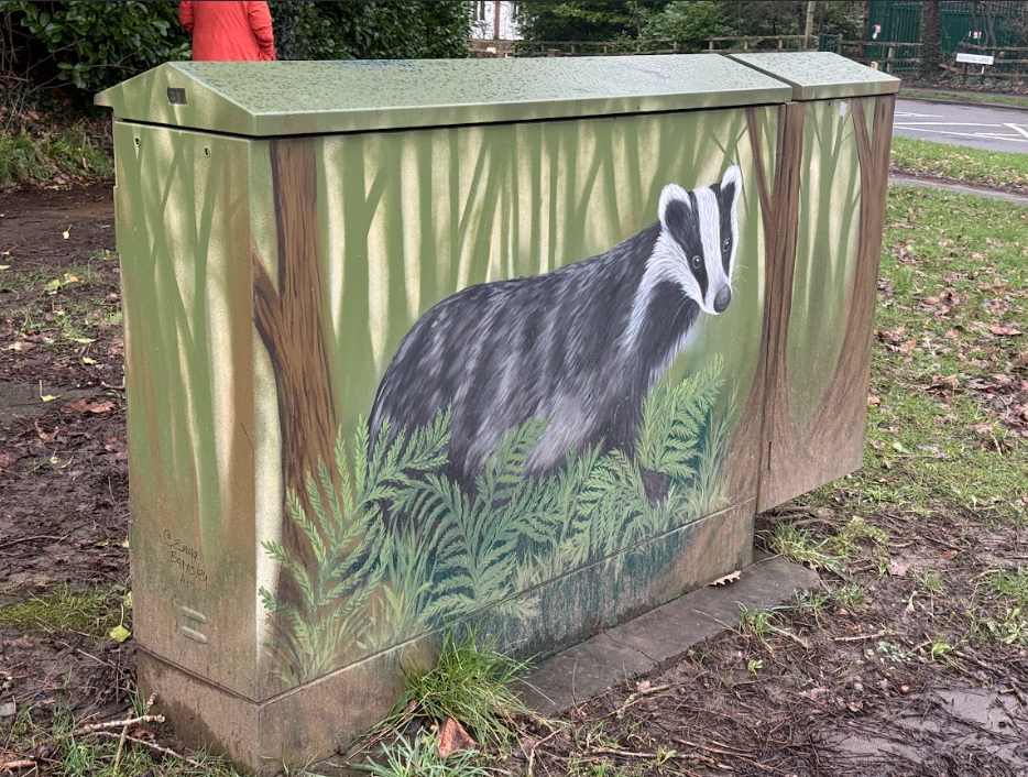 A green utility box with a badger painted on it, in a woodland scene.