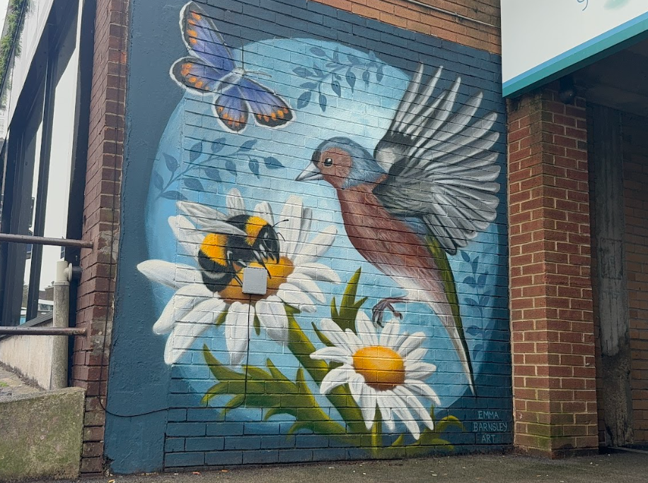 Emma's winning mural on the side of a building. A painted image of a bird, two daisies, a bee on one of the daisies, a butterfly, and some plants on a blue background.
