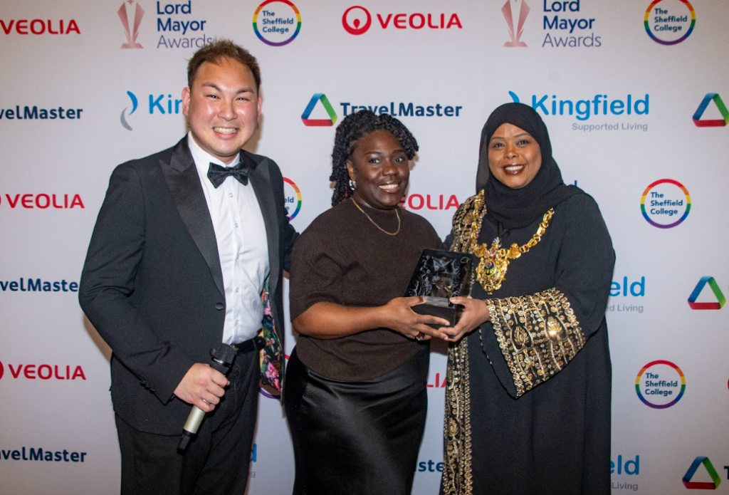 Presenter, award winners and Lord Mayor, stood in that order. Winner holding the Lord Mayor award. All smiling. 