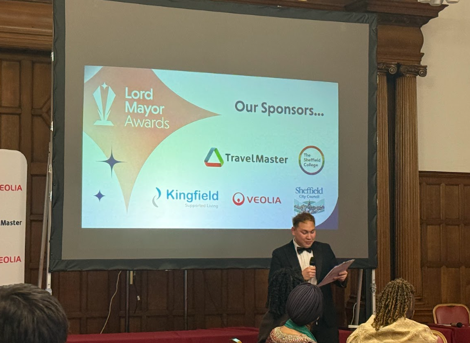 Presenter of the Lord Mayor awards stood in front of a screen, with the event sponsor logos on. 