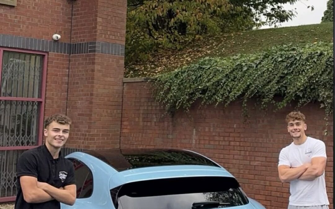 Two adults standing with their arms crossed smiling infront of the rear-side of a car