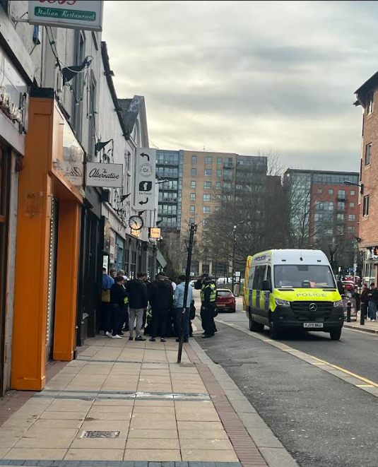 A shot from further down the street showing a larger group of men involved in a conflict with one another outside of a sports bar whilst police intervene, there is one police van in the back ad onlookers watching