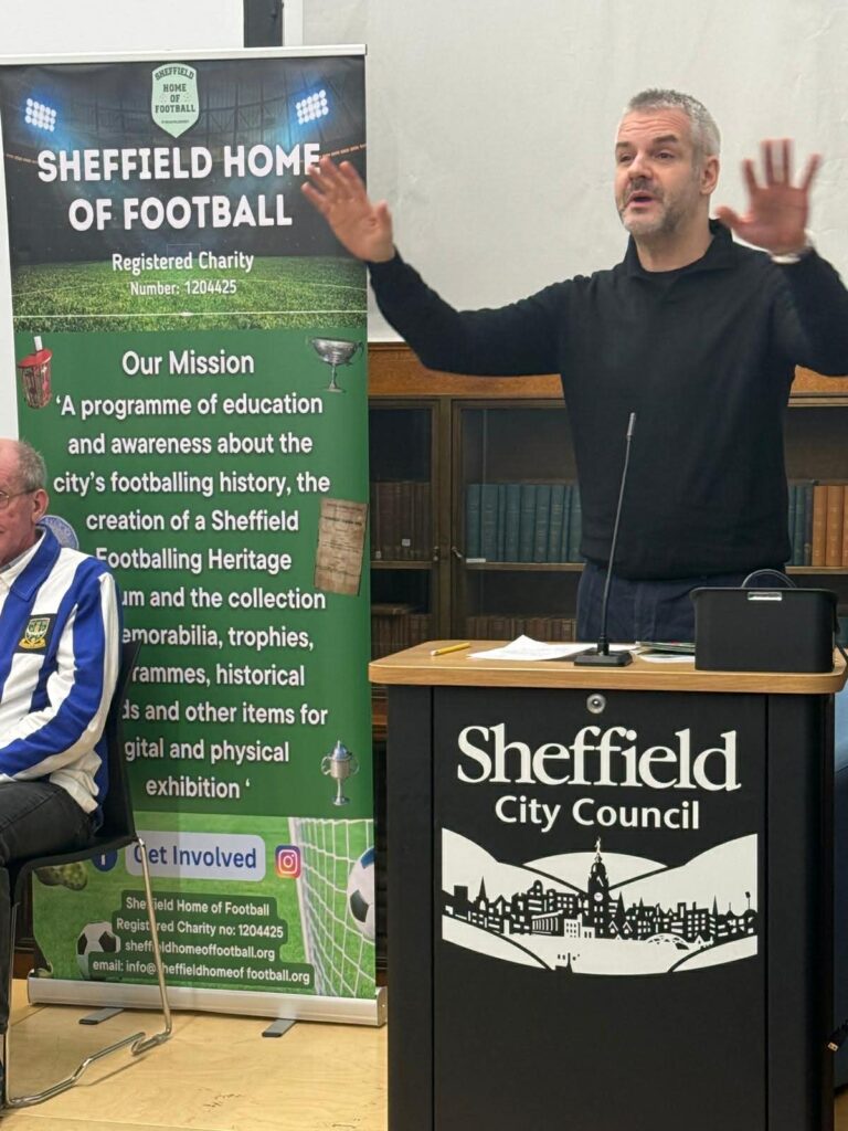 A man giving a talk with a Sheffield Home of Football banner behind him