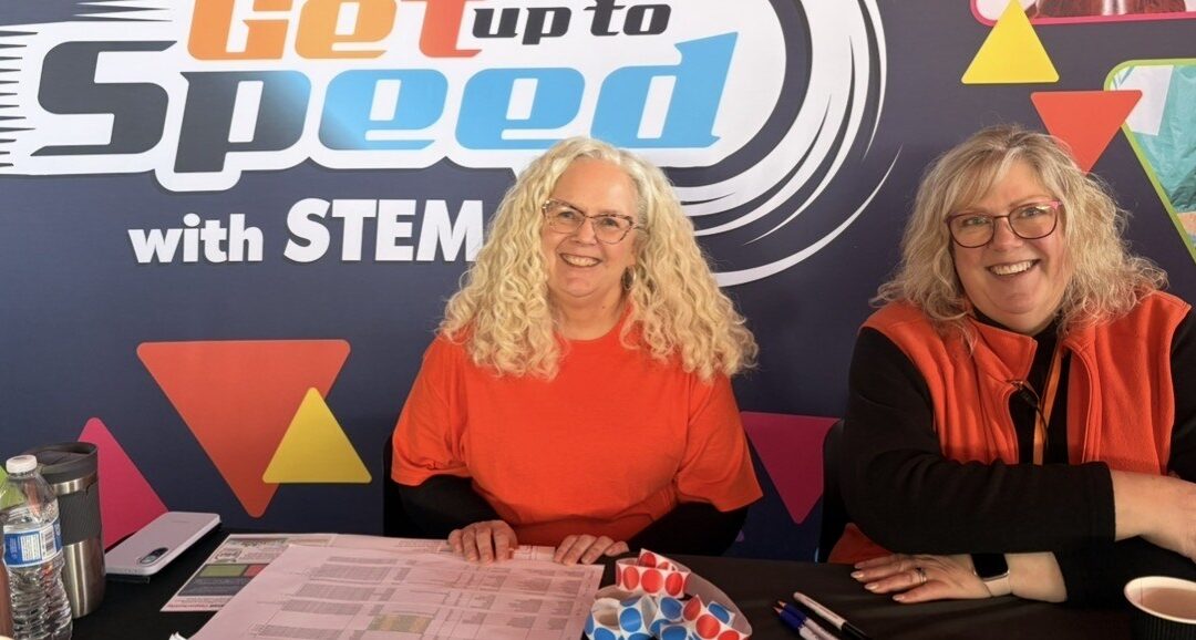 Two women in orange shirts in front of the Get up to Speed with STEM banner