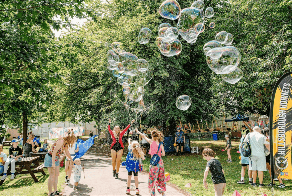 Kids and adults on a path surrounded by trees, at Hillsborough park, smiling, playing with large bubbles.