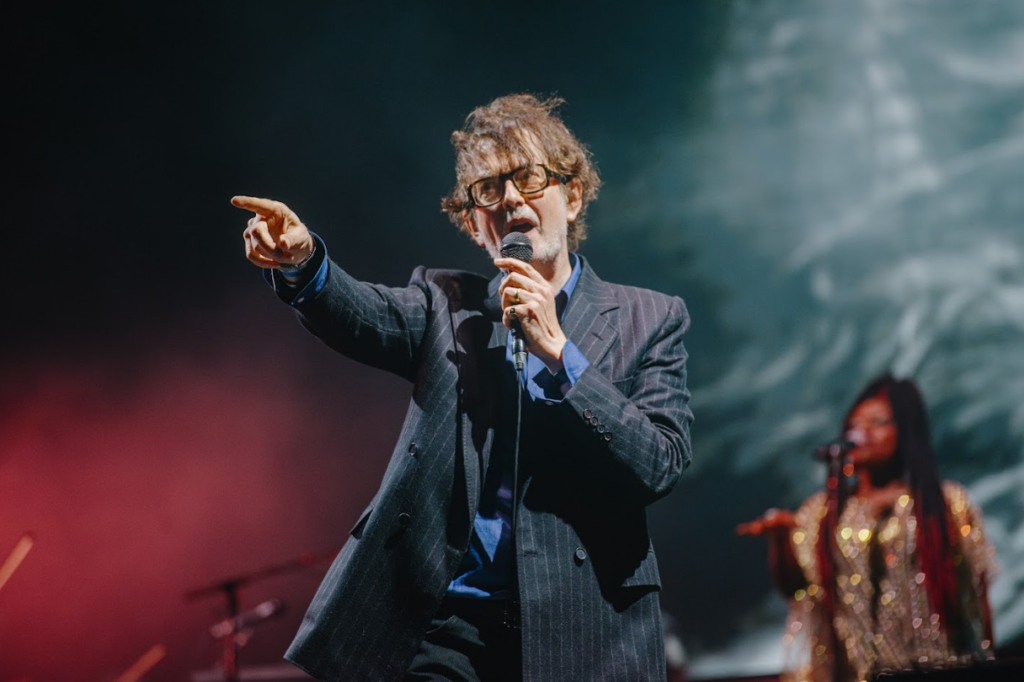 Jarvis Cocker, of Pulp, on stage, pointing out at the crowd. 