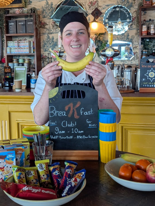 woman in a chef hat holding up a banana behind a sign that says "Breakfast club 9am- 10am" 