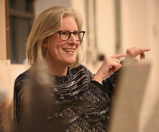 Image of Esther Richardson smiling and pointing, in a beige coloured room. While wearing woolen jumper.