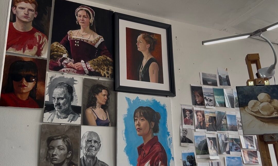 A wall of paintings of people, with a painting of lemons on an easel.
