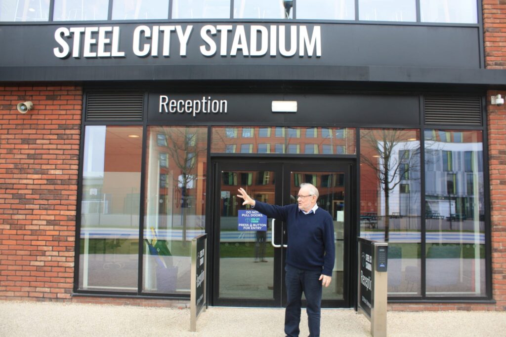 Photograph of Richard Caborn in blue jumper outside the Steel City Stadium pointing.