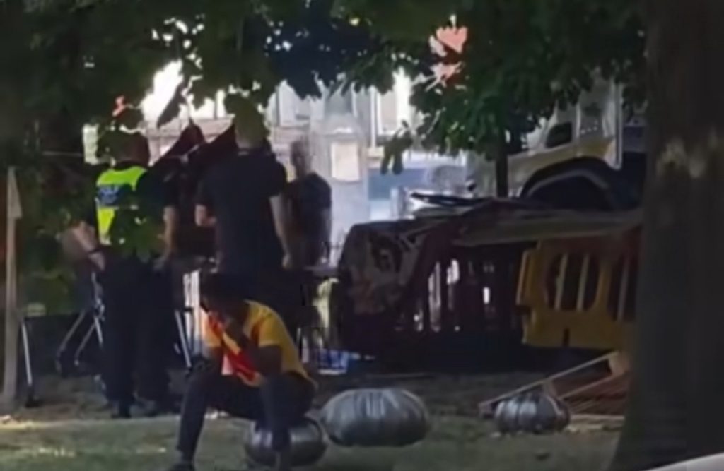 Police and Council workers remove the homeless camp in the background, in the foreground a man sits on a seat in the park.